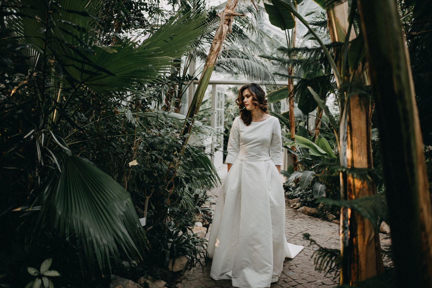 Bride in white gown walking through lush tropical greenhouse surrounded by palm leaves and green plants.