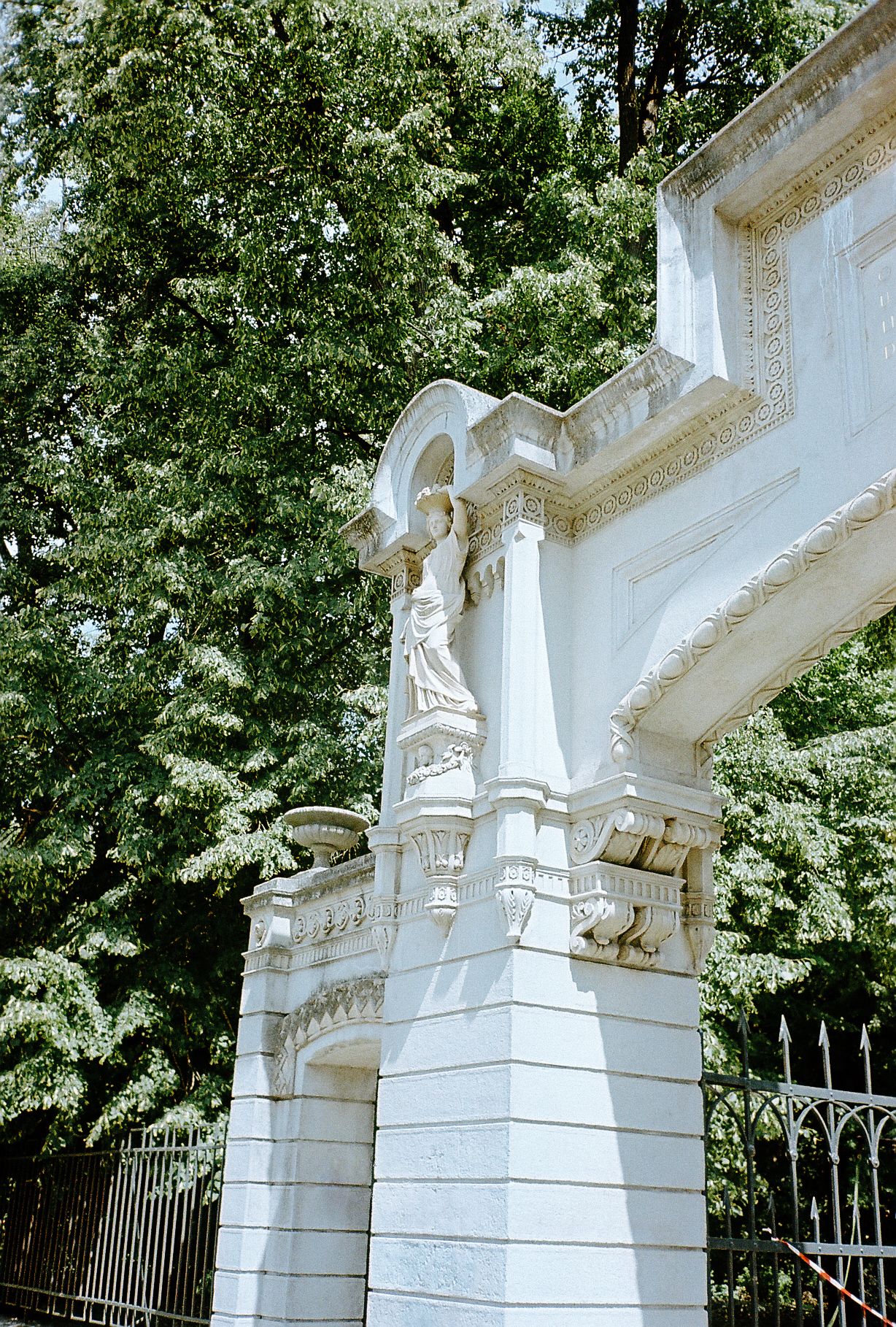 White marble archway with ornate architectural details and statues stands against a backdrop of green trees.
