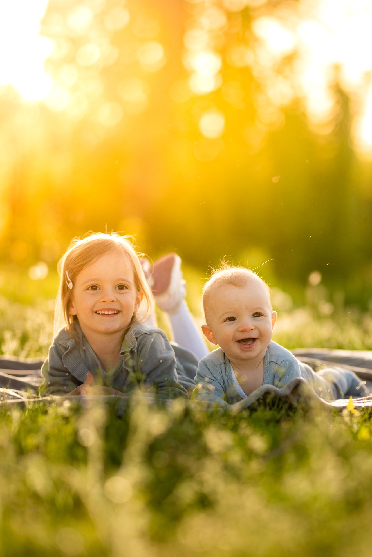 Two happy children lying on a blanket in a sunny green meadow, smiling joyfully in golden hour light.
