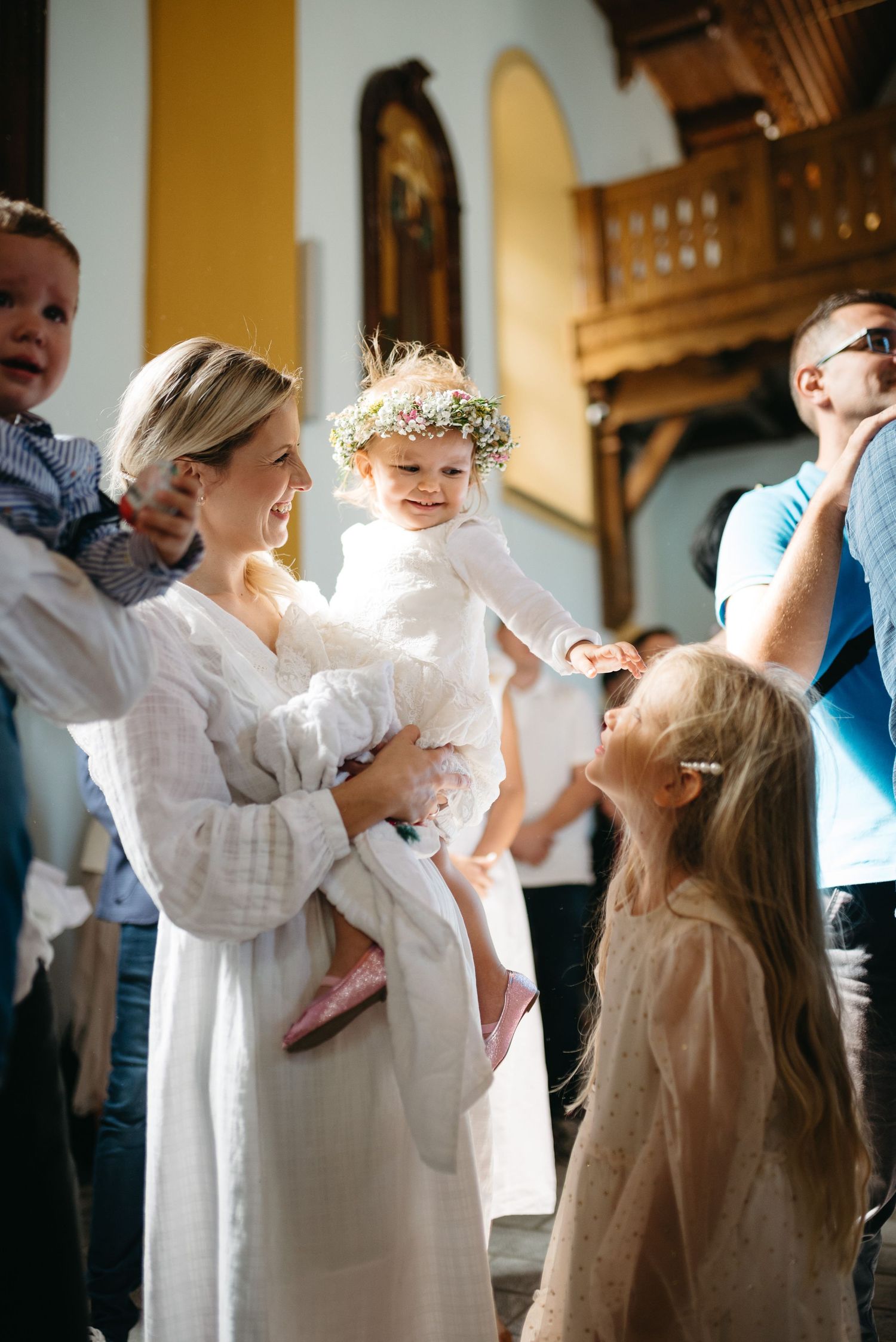 Woman in white dress holds toddler with flower crown at a church celebration with children and guests.