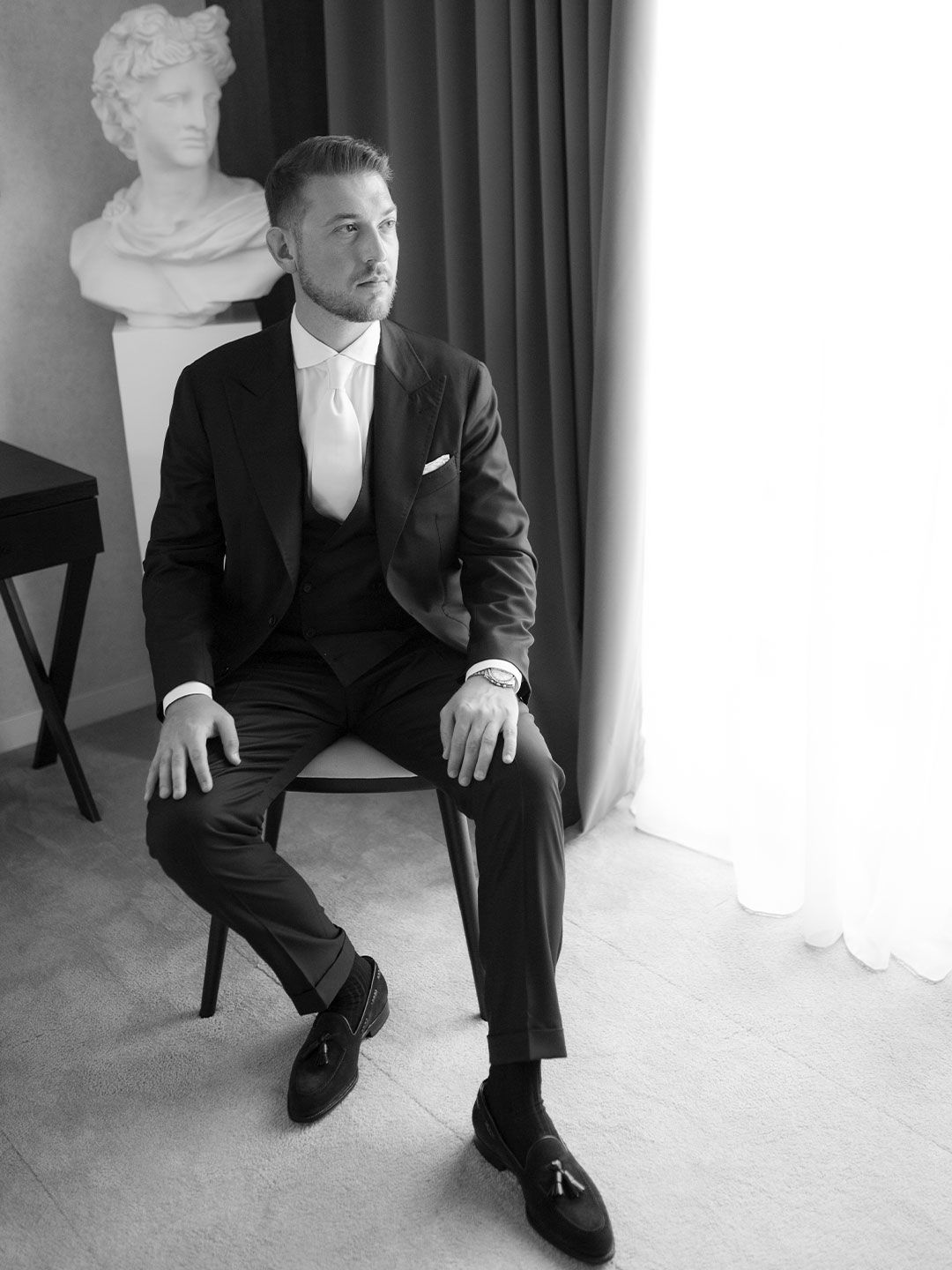 A well-dressed man in a dark suit sits confidently on a chair beside a classical bust sculpture in a black and white photo.