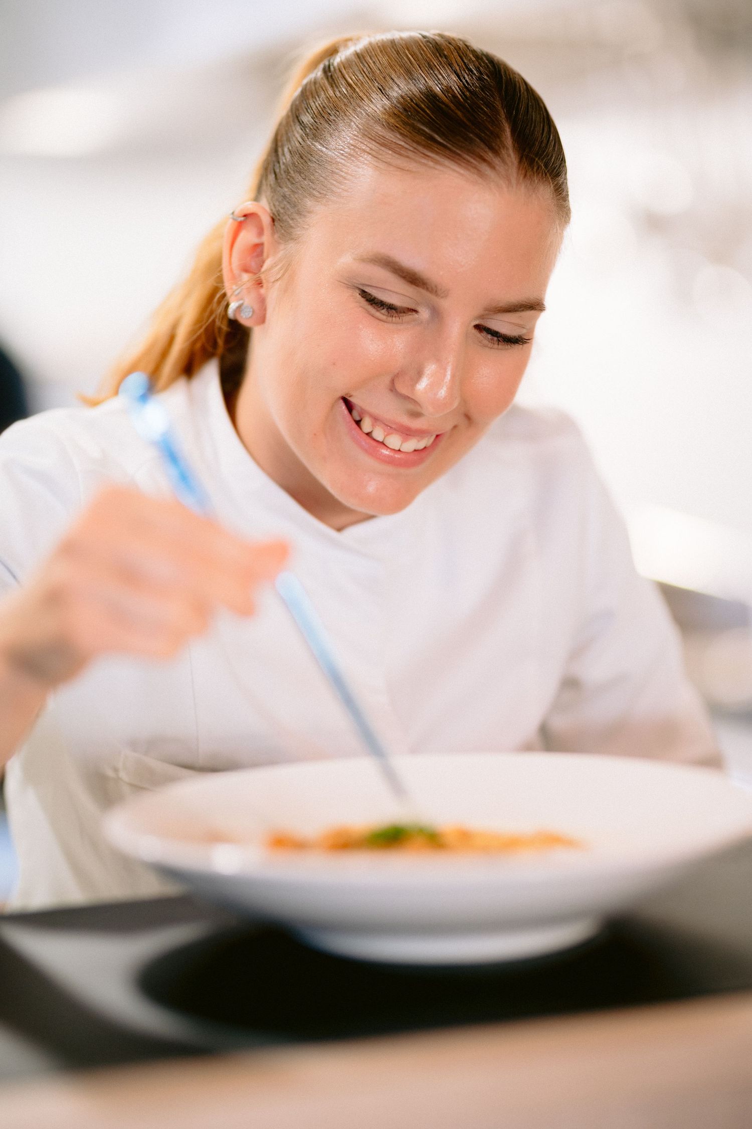 Smiling female chef carefully garnishing a bowl of soup with a blue utensil in a professional kitchen.