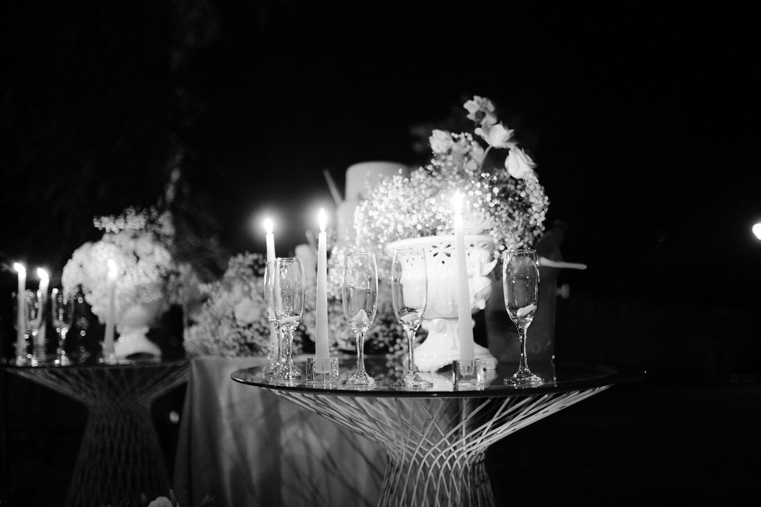 Black and white photo of an elegant candlelit table display with floral arrangements and glassware at a nighttime event.