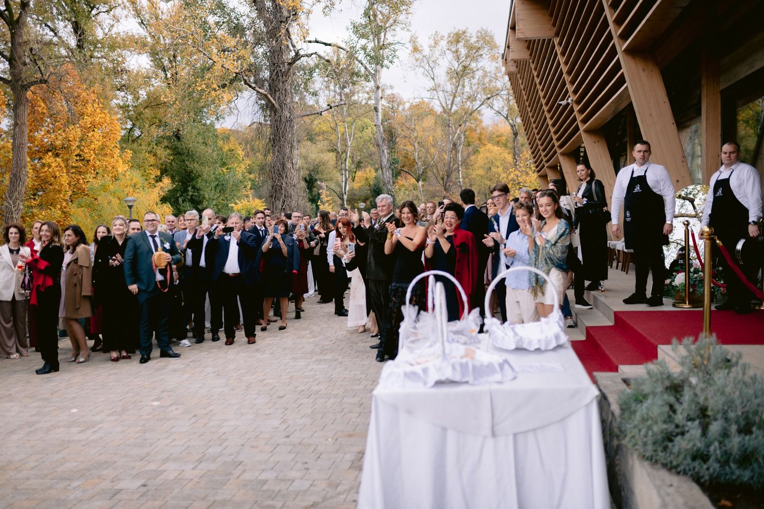 Outdoor wedding reception with guests gathered on a red carpet pathway surrounded by autumn trees.