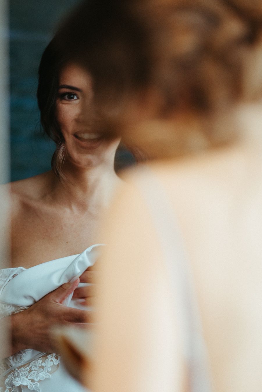 Smiling bride in white lace wedding dress adjusting her gown while looking in a mirror on her wedding day.