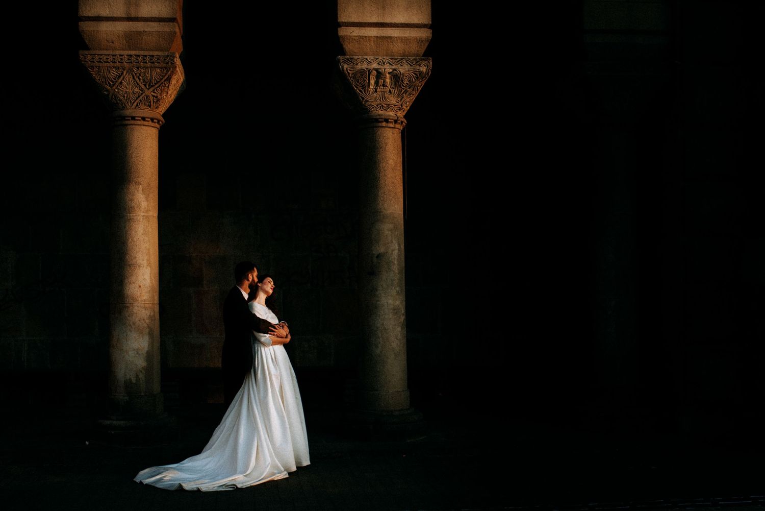 Bride and groom embracing in dramatic light between ancient stone columns at a historic wedding venue.