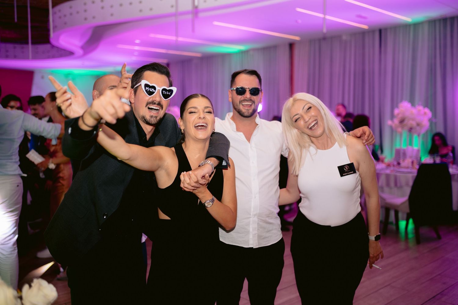 Wedding guests dance and celebrate on a dance floor illuminated by purple and blue lighting at an indoor reception venue.