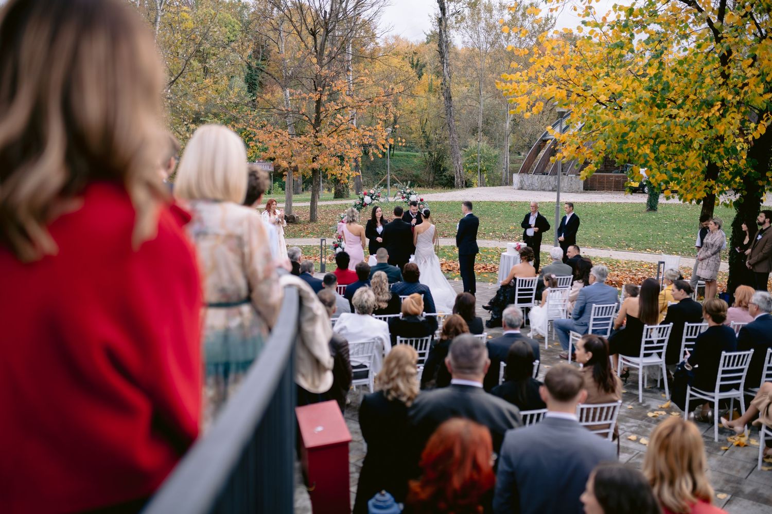 Autumn outdoor wedding ceremony with guests seated on white chairs among fall foliage.