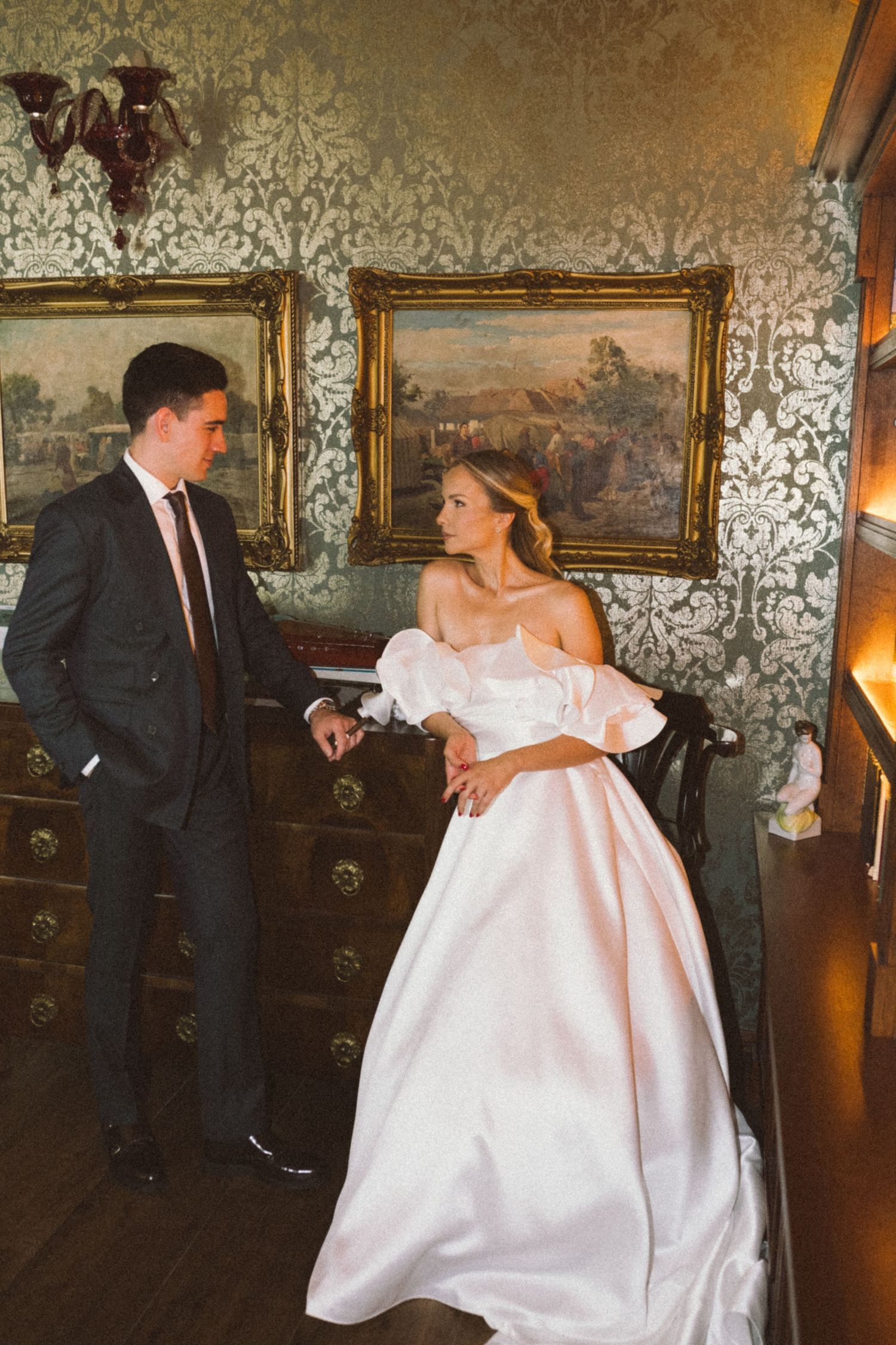 A couple in formal wedding attire stands in an elegant room with ornate wallpaper and framed landscape paintings.