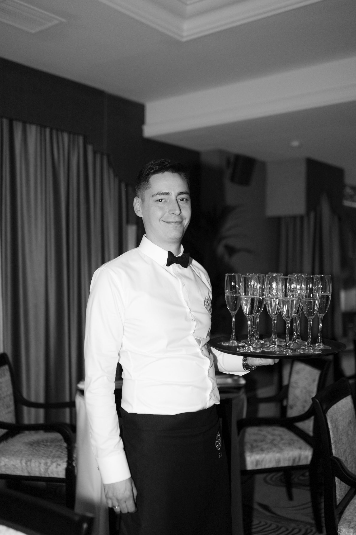 Professional waiter in bow tie holding tray of champagne flutes in black and white restaurant photo.