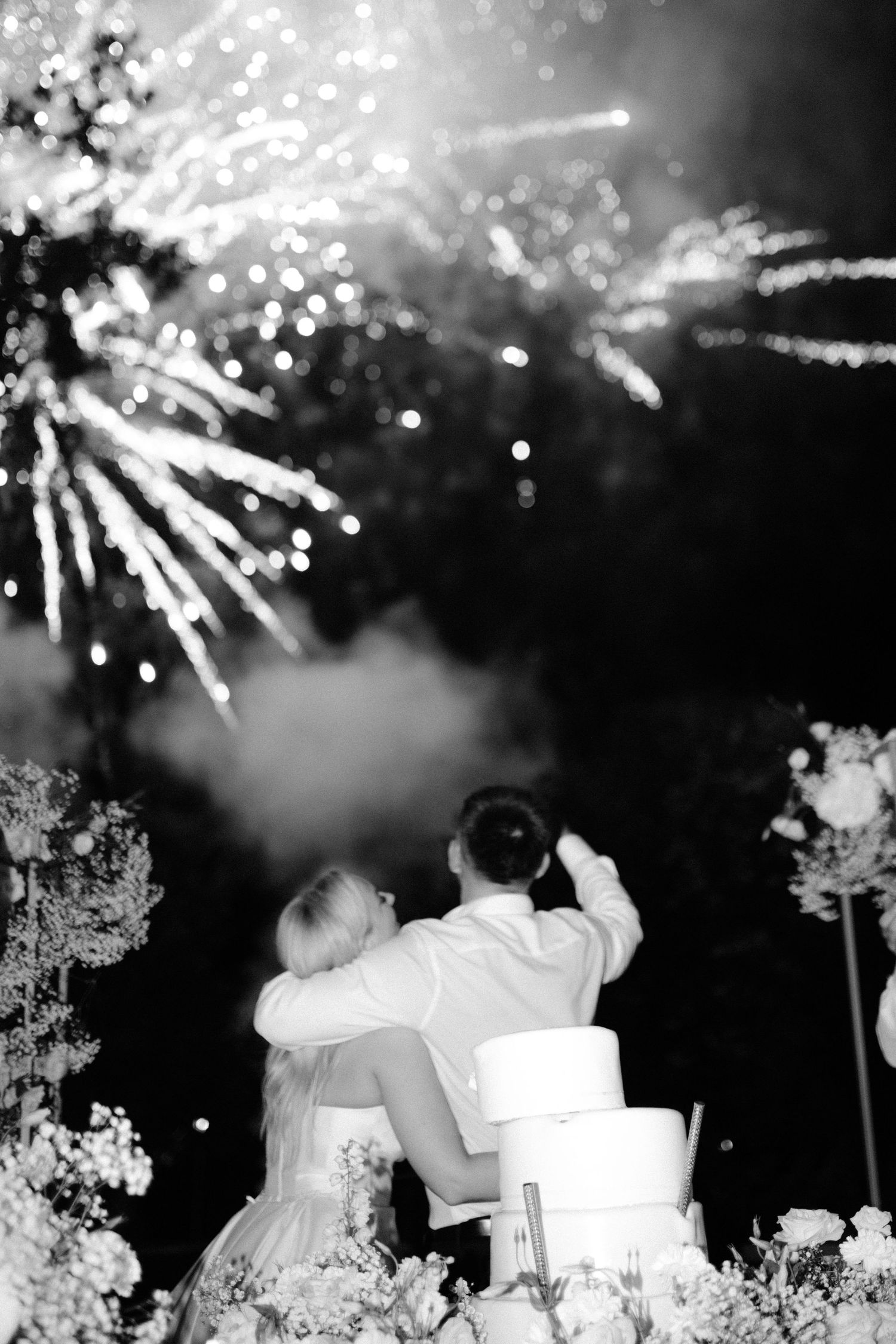 Couple watching fireworks display next to their wedding cake in black and white.