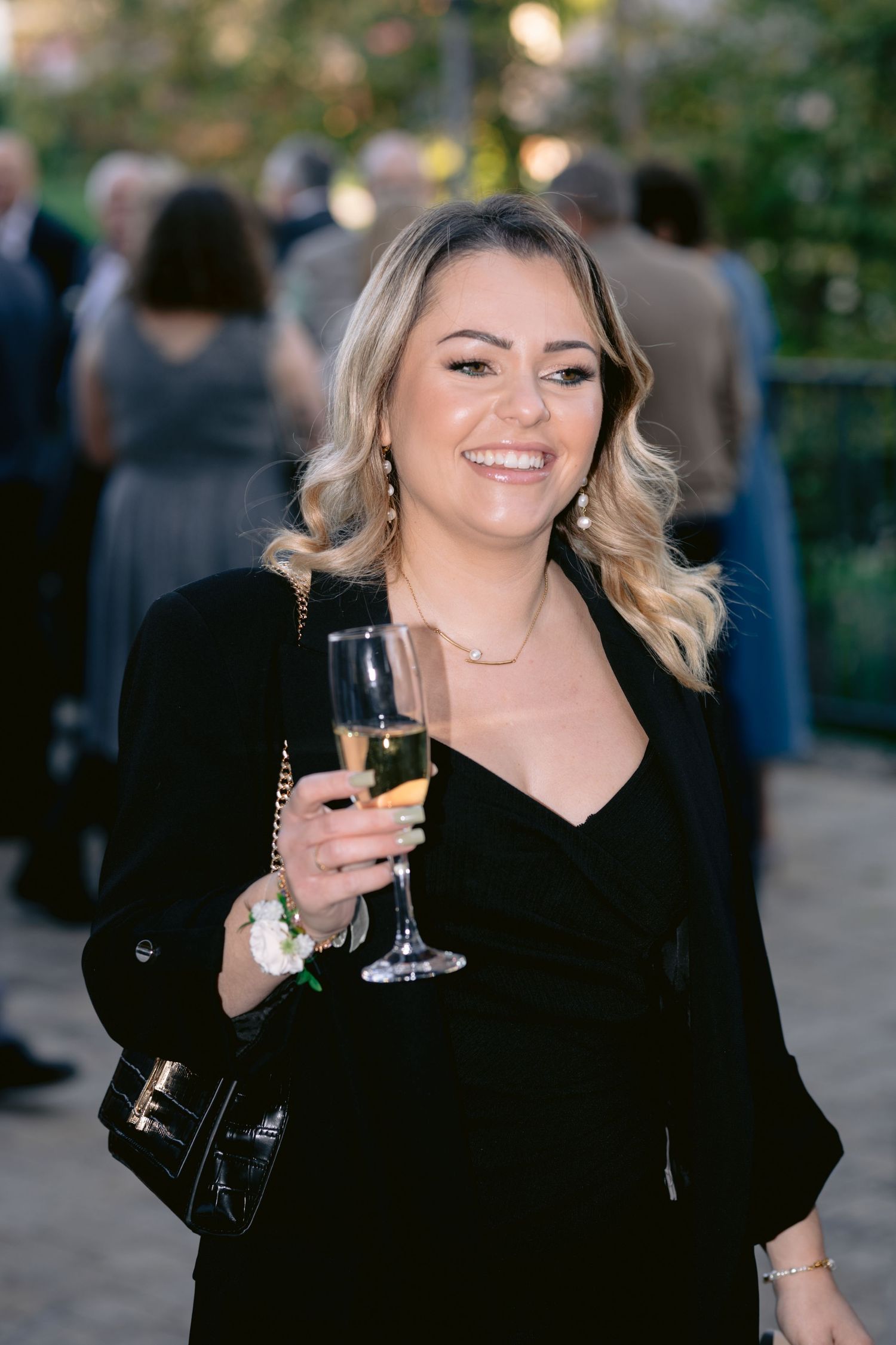 Elegant woman in black dress holding champagne glass at outdoor evening event with blurred background.