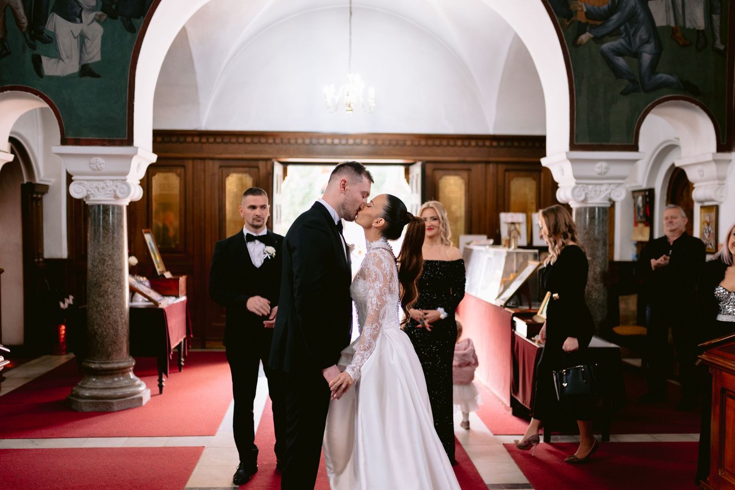 A wedding ceremony takes place inside a traditional church with white arches and red carpet.