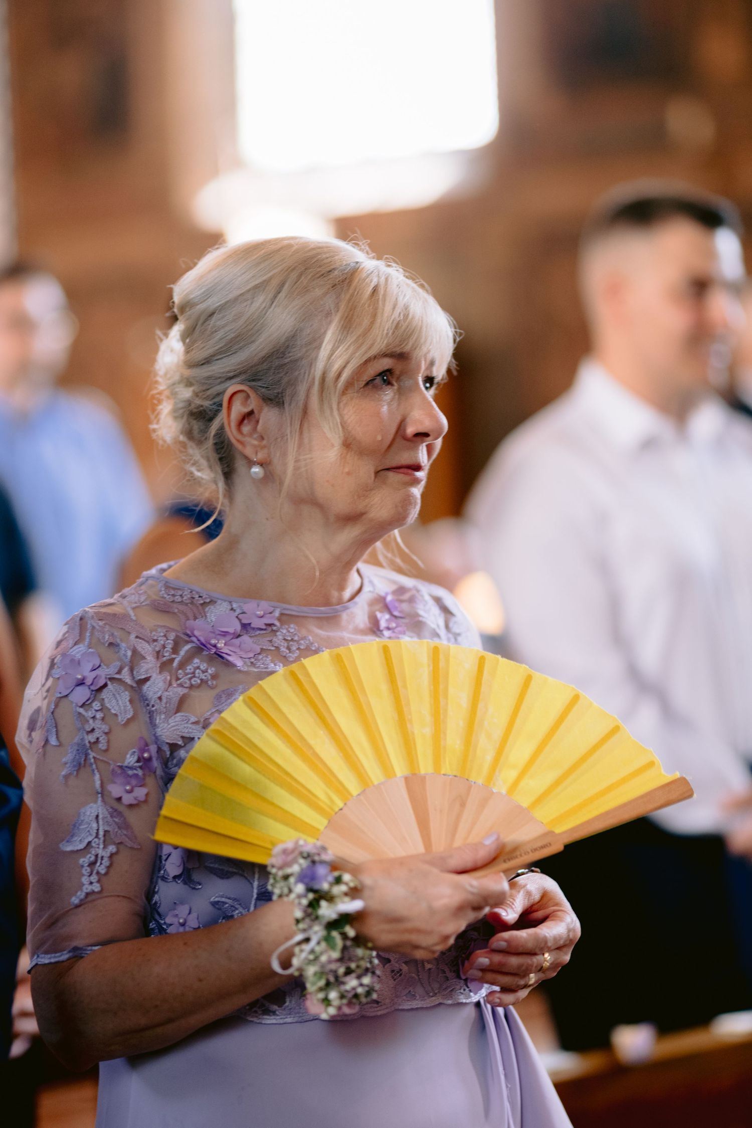 Elegant senior woman in lavender lace dress holding yellow hand fan at a wedding ceremony.