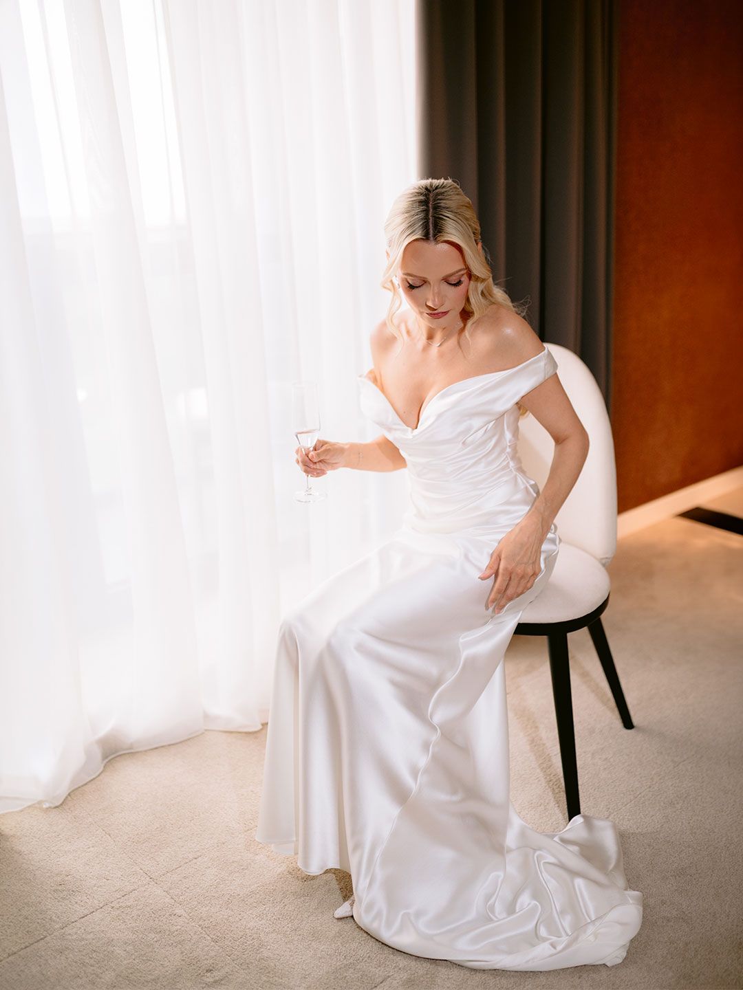 Elegant bride in off-shoulder white satin wedding gown seated in chair holding champagne glass by sheer curtains.