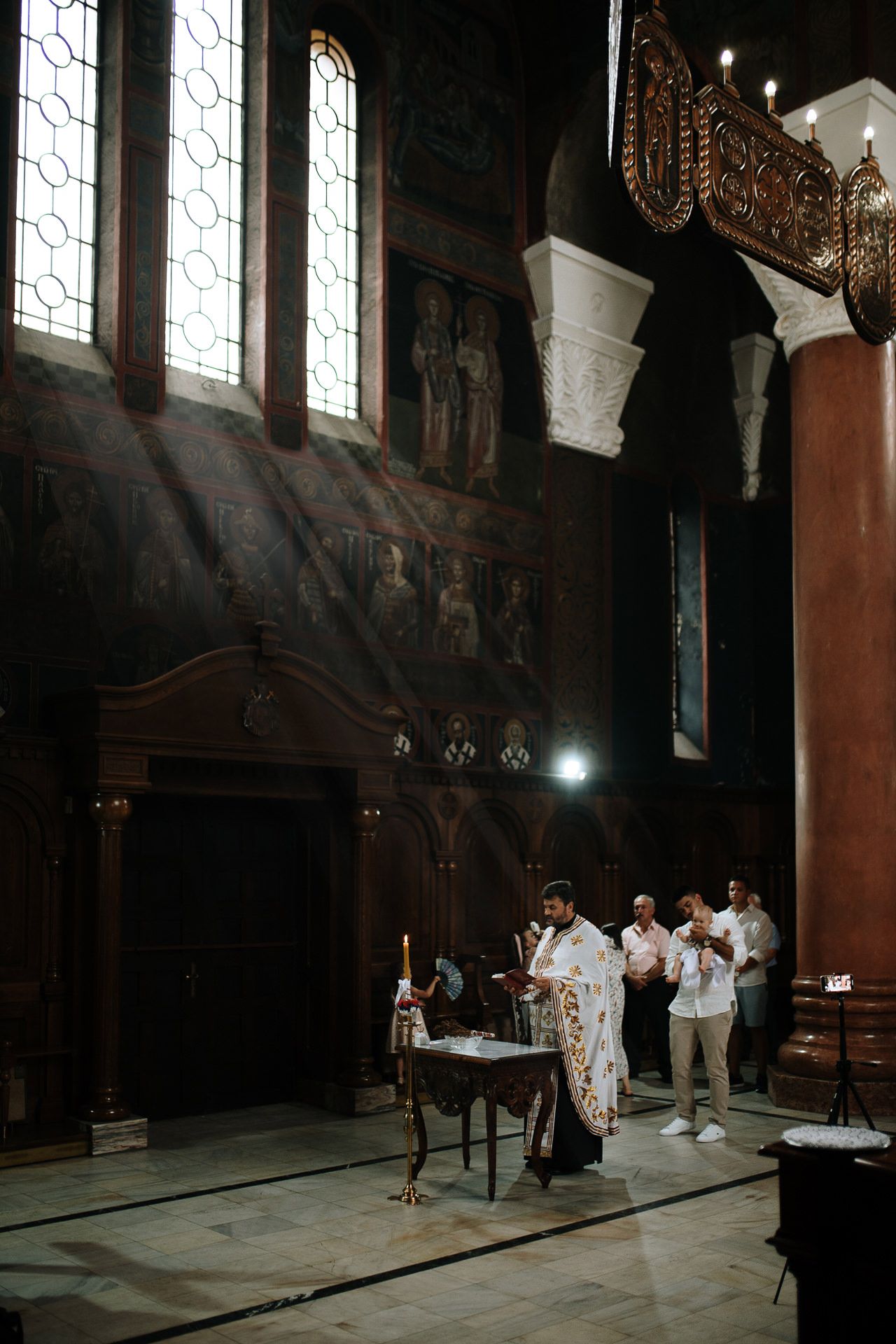 Priest performing a religious ceremony inside an ornate Orthodox church with dramatic light rays through stained glass windows.