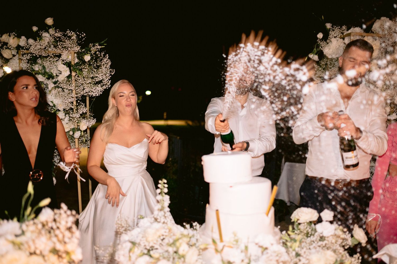 Bride in white dress laughing near tiered wedding cake with sparklers and champagne at outdoor night reception.