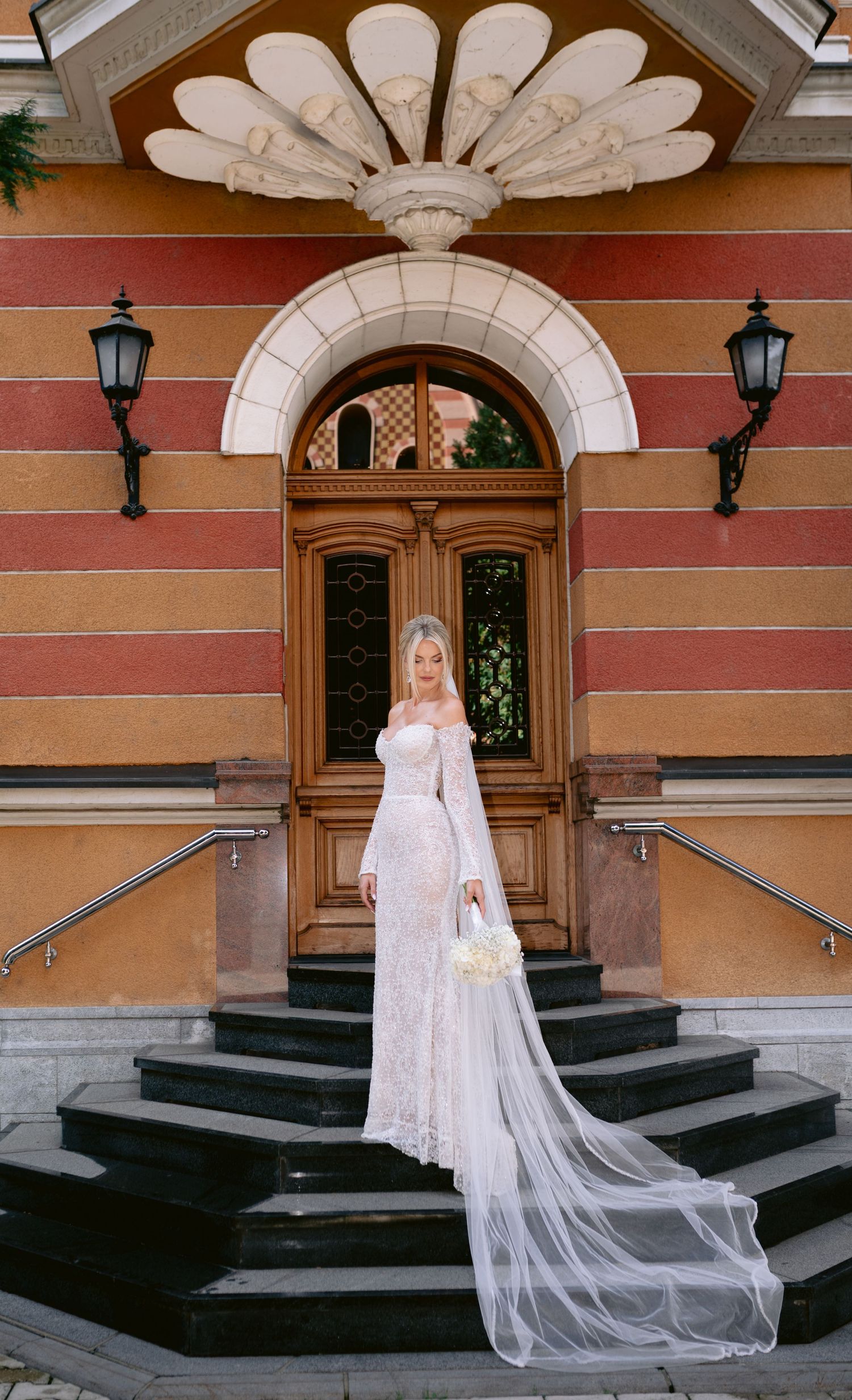 Bride in flowing white dress and veil poses on grand building steps with ornate architecture.