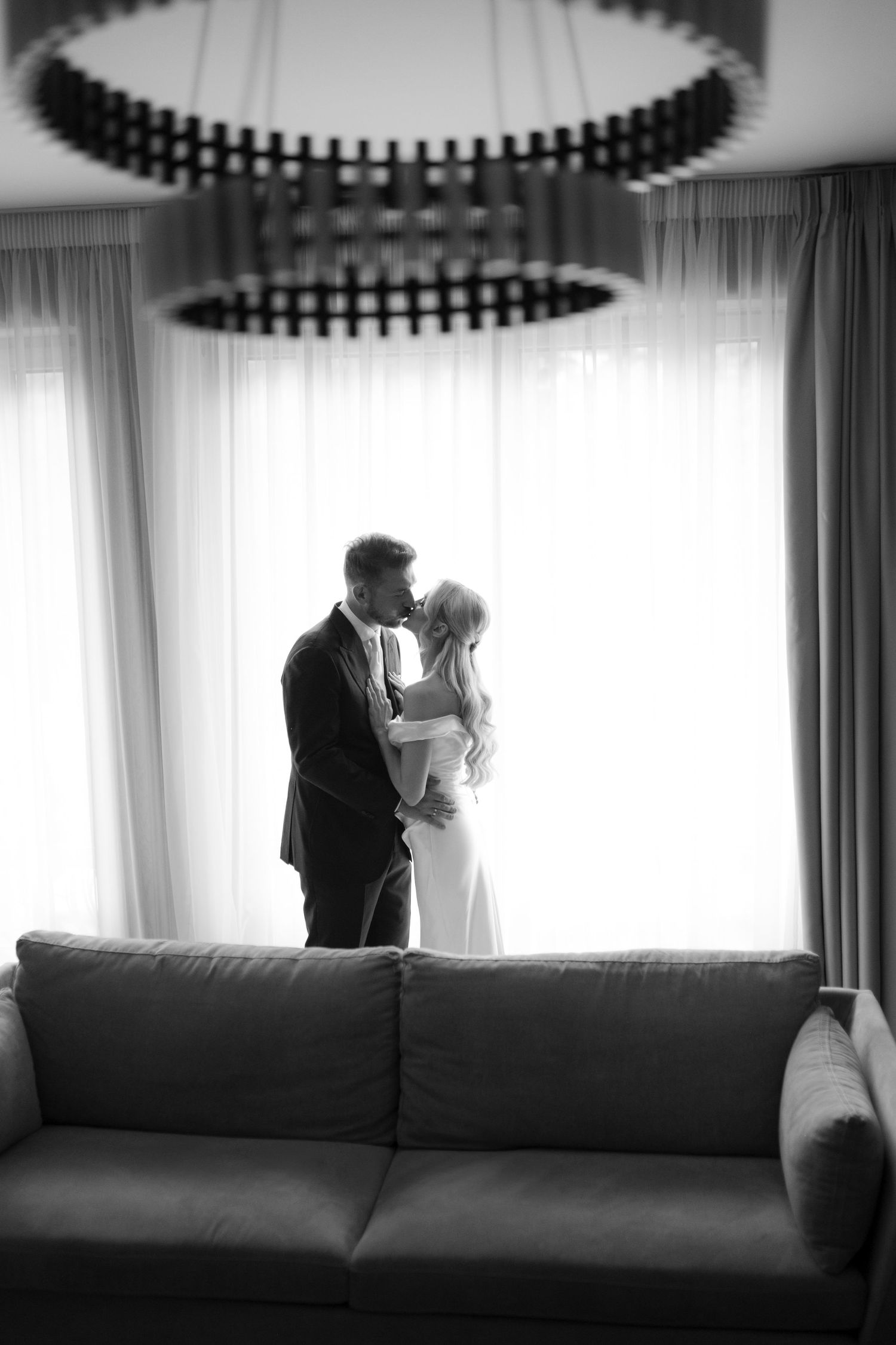 Crno-bijela fotografija mladenaca u elegantnoj vjenčanoj odjeći dok se grle i ljube pored svijetlog prozora u enterijeru