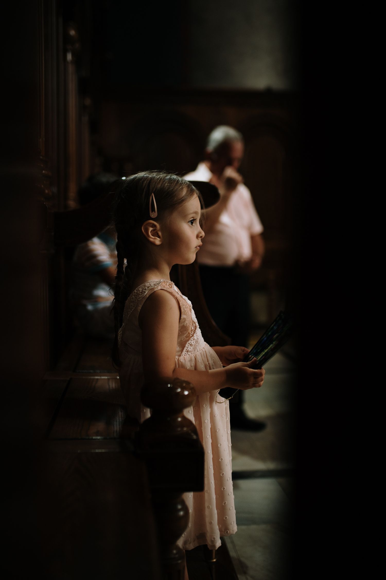 Young girl in pink dress holding a book, standing attentively in a dimly lit church interior.
