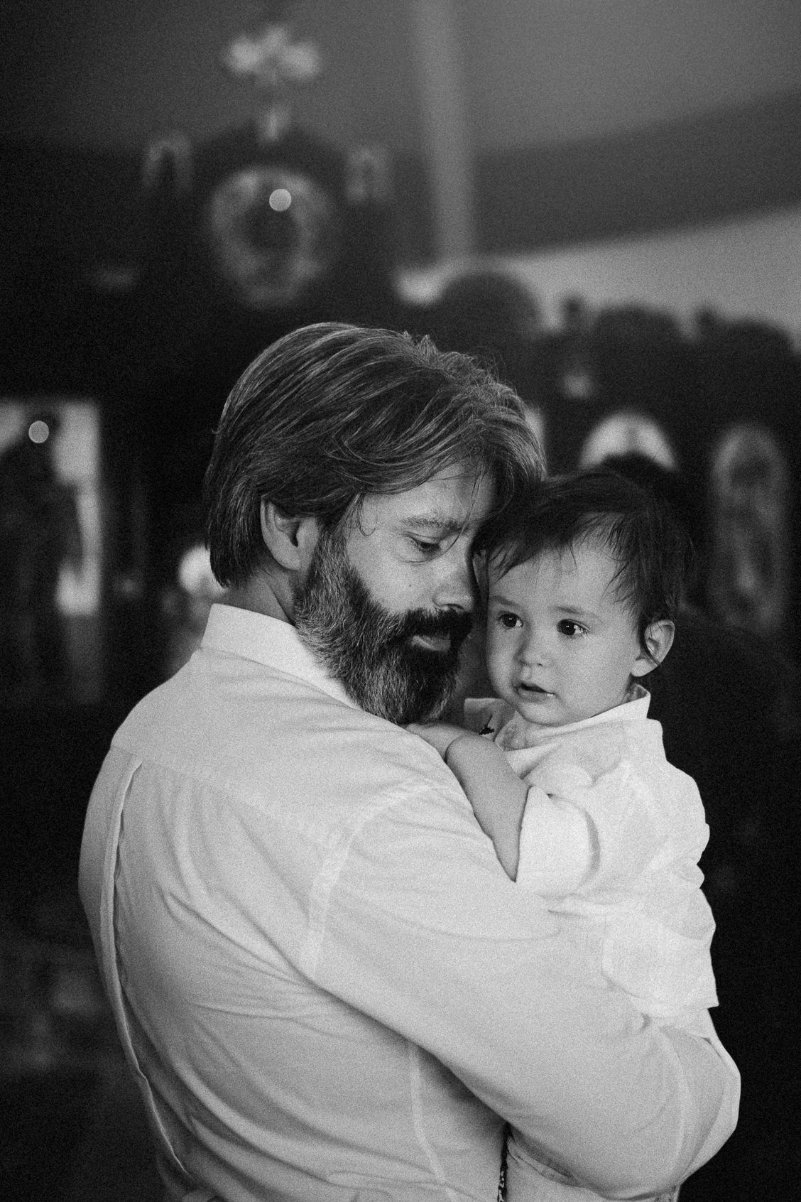 Black and white photo of a bearded man tenderly holding a baby in his arms indoors.