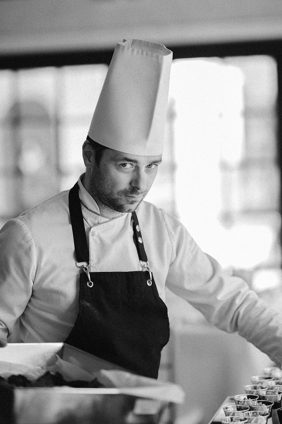 A chef in white uniform and black apron works intently in a professional kitchen, wearing a traditional tall toque.