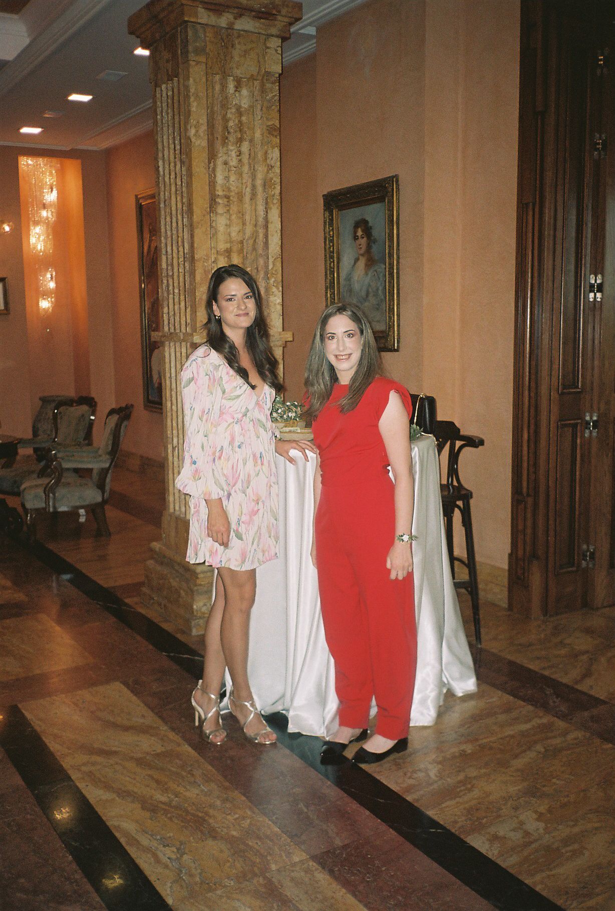 Two people in contrasting outfits, one in pink floral and one in red, stand in an elegant hallway with marble floors.