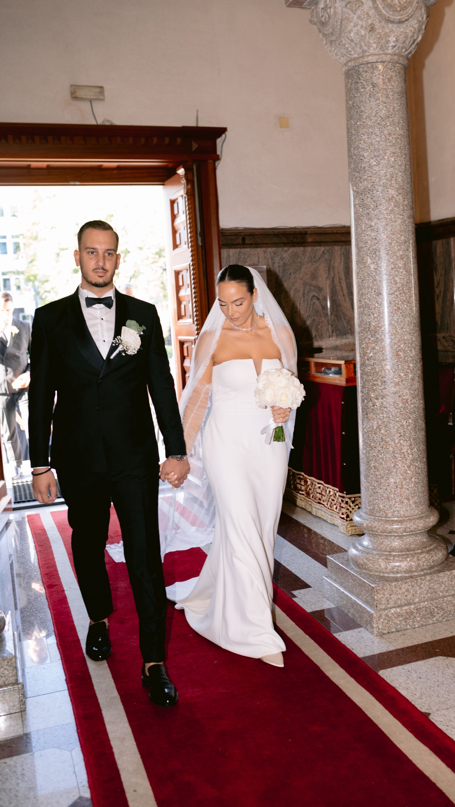 A bride in a white strapless gown and groom in a black tuxedo walk down a red-carpeted aisle in an ornate venue.
