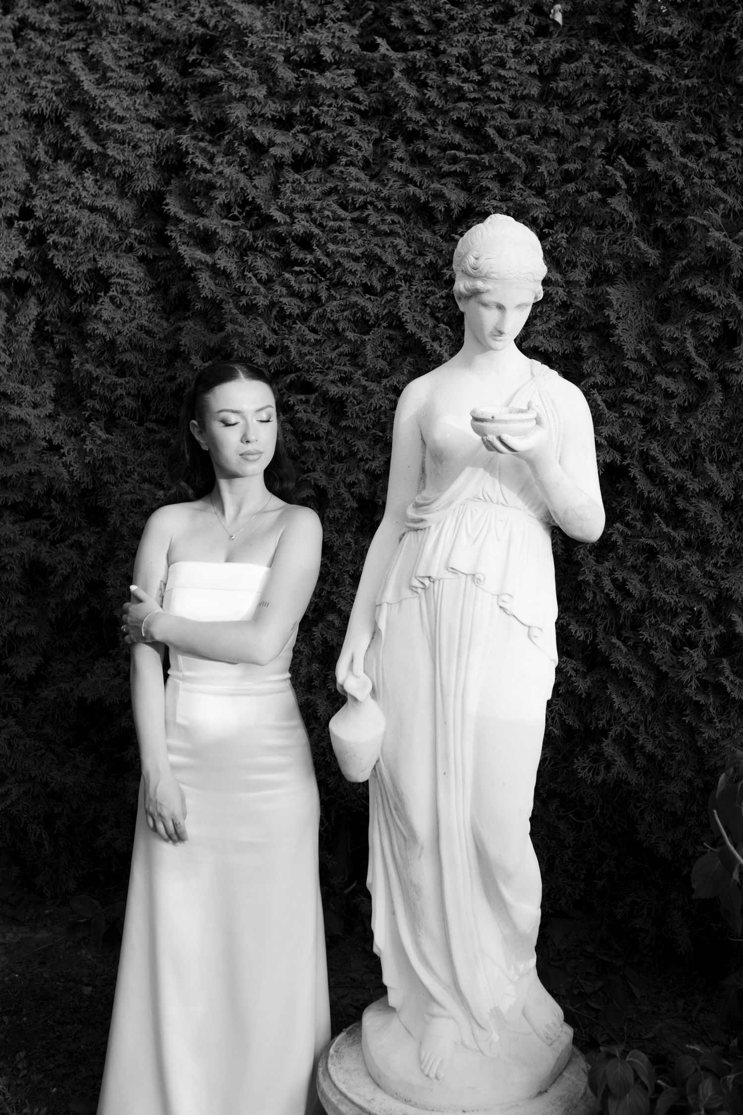 Black and white photo of a woman in a white gown standing beside a classical female statue in a garden.