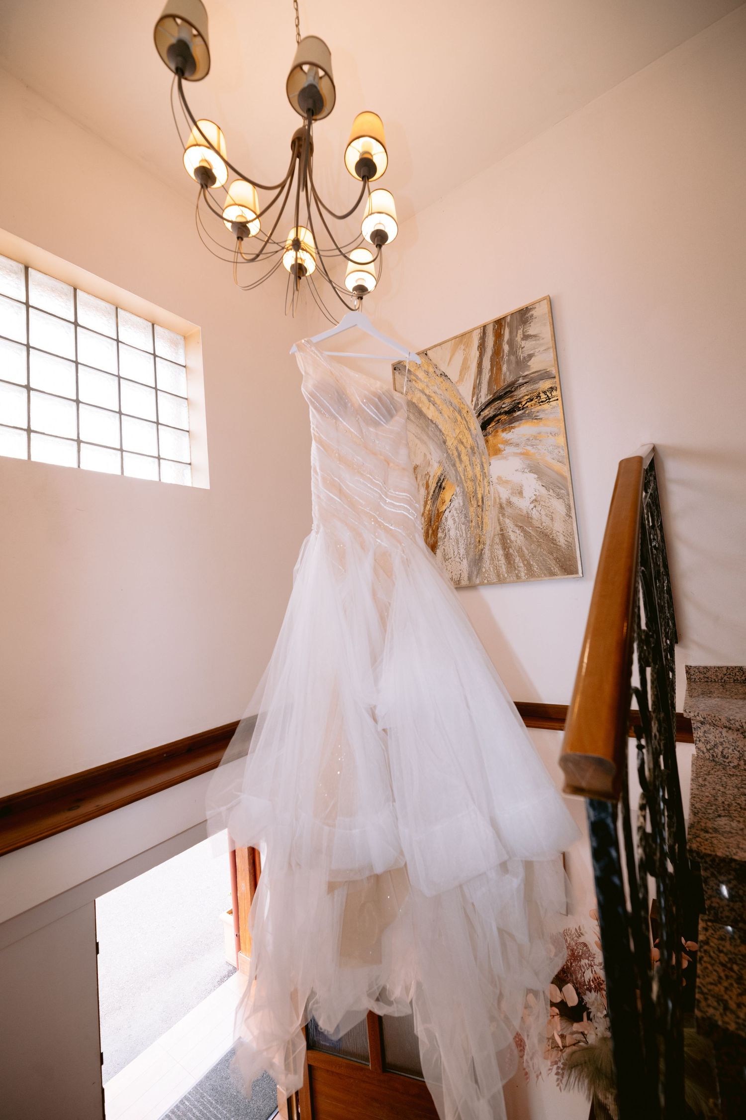 White wedding dress hanging from chandelier in elegant stairwell with decorative painting.