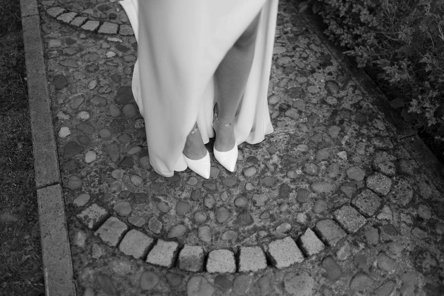 Black and white photo of a bride in white heels standing on a cobblestone path in a wedding gown.