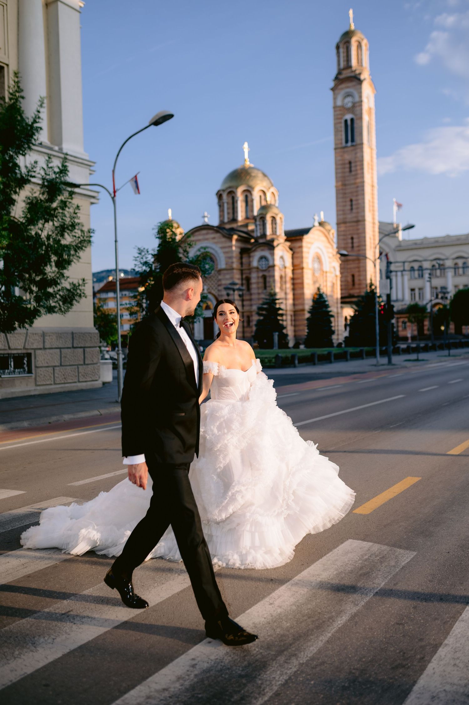 Bride and groom crossing a city street in formal wedding attire with a historic church tower in the background.