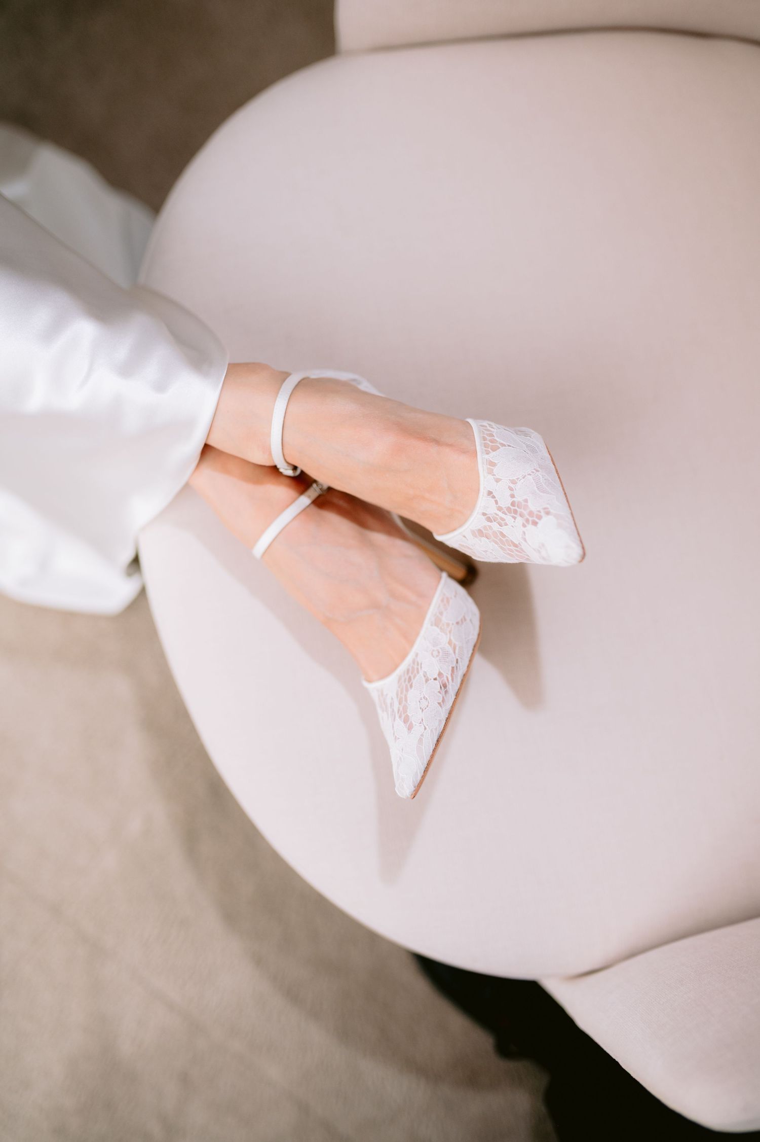 Elegant white lace bridal heels with ankle strap on a cream chair, perfect wedding shoes for brides.