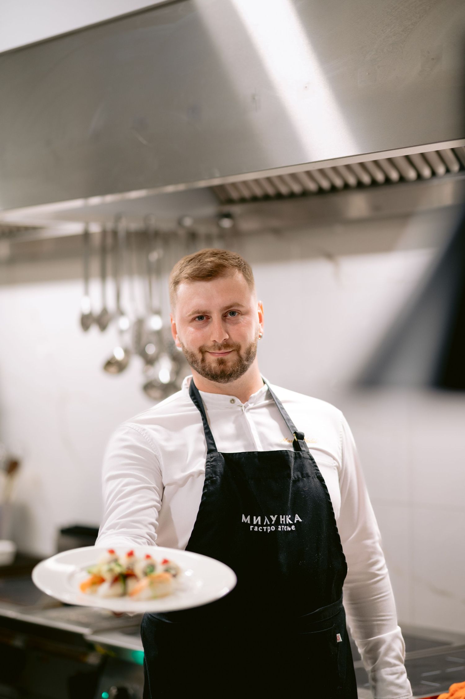 Smiling male chef in black apron holding a plate of gourmet food in a professional restaurant kitchen.
