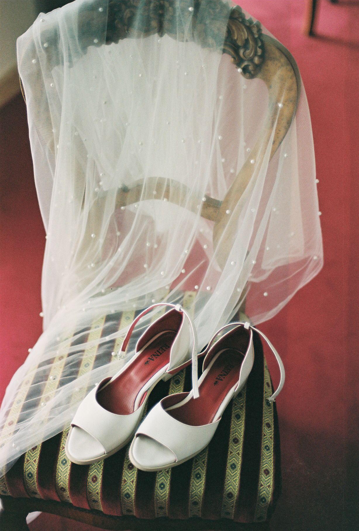 A row of white bridal shoes lined up against a red background with flowing tulle veil draped over them.