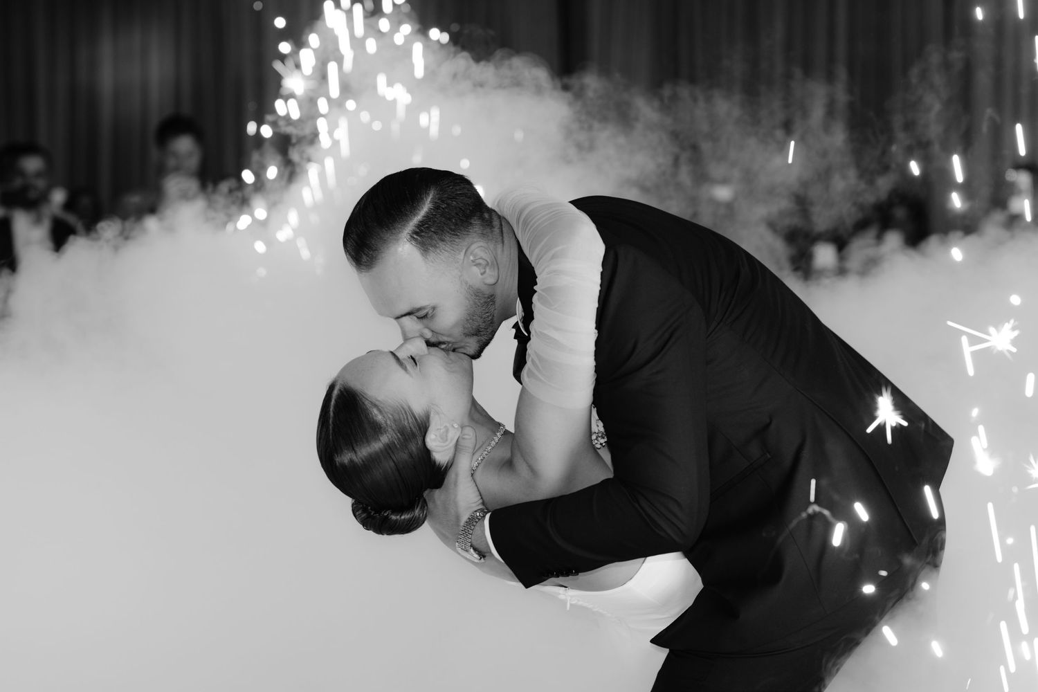 A couple in black attire performs an intimate dance move in front of a light-filled backdrop.
