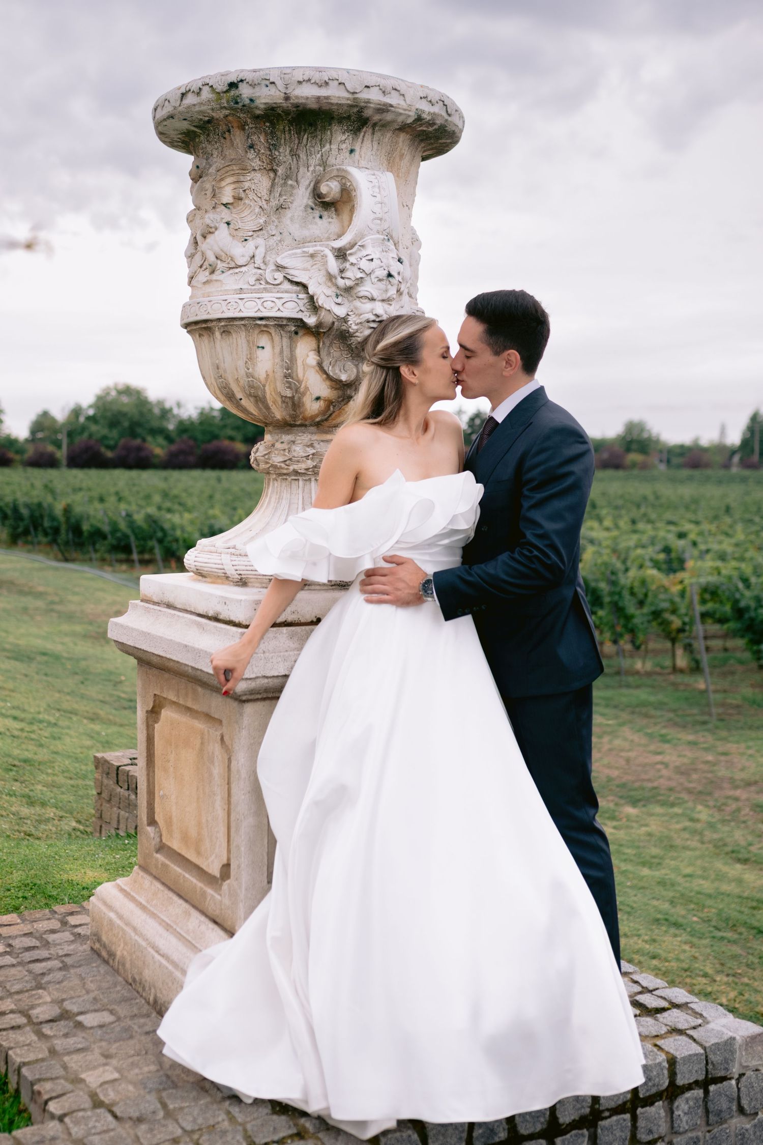 Romantic wedding couple embracing by ornate stone pillar in vineyard setting.