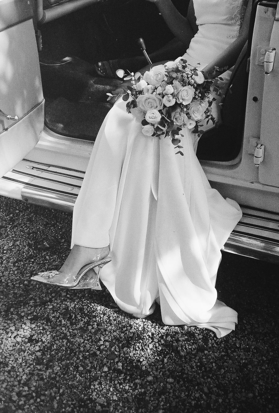 Black and white photo of a bride in a white gown holding a floral bouquet, stepping out of a vintage car in heels.