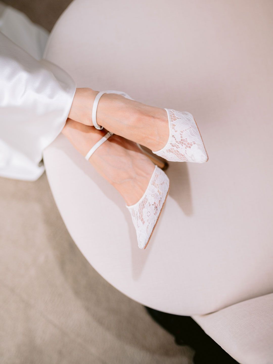 White lace pointed-toe heels with ankle strap resting on a beige surface, perfect bridal wedding shoes.