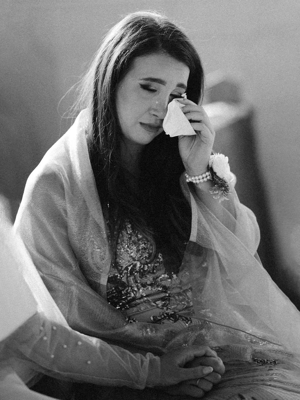 Black and white photo of a young woman in traditional attire wiping tears with a tissue, wearing pearl bracelet.