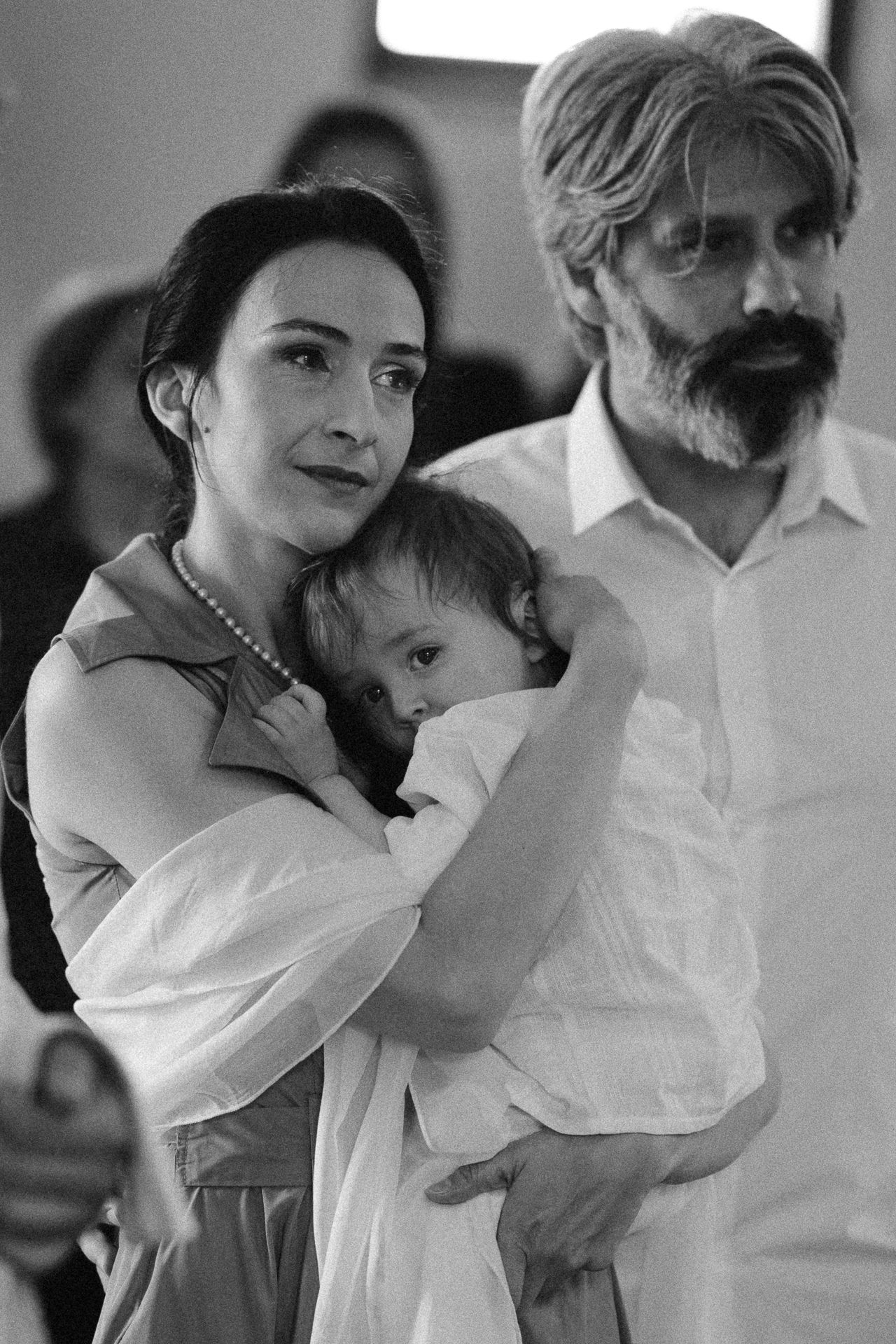 Black and white photo of a woman holding a baby with a bearded man standing behind her at a family gathering.