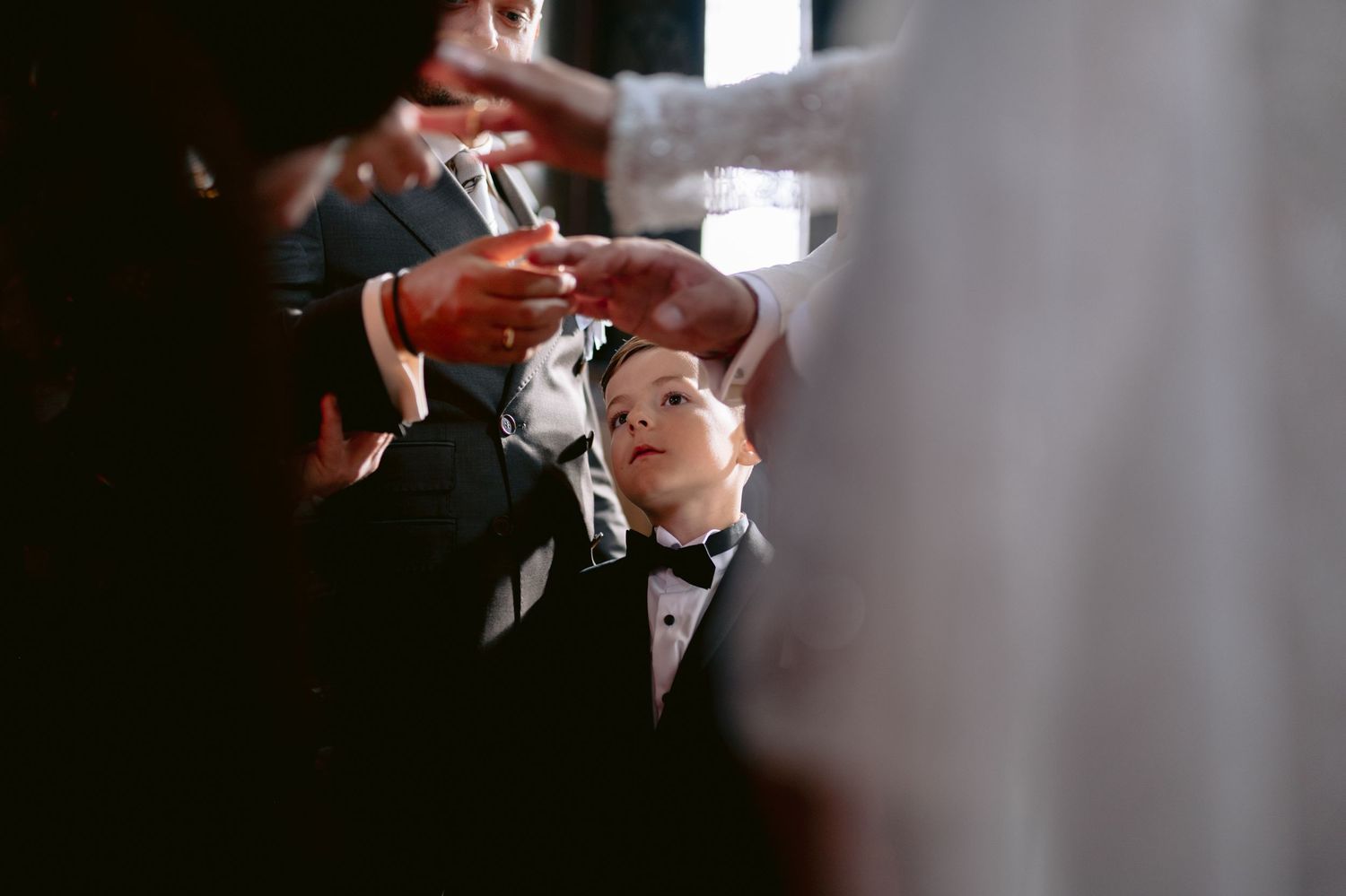 A young boy in a tuxedo looks up at adults exchanging rings during a wedding ceremony.