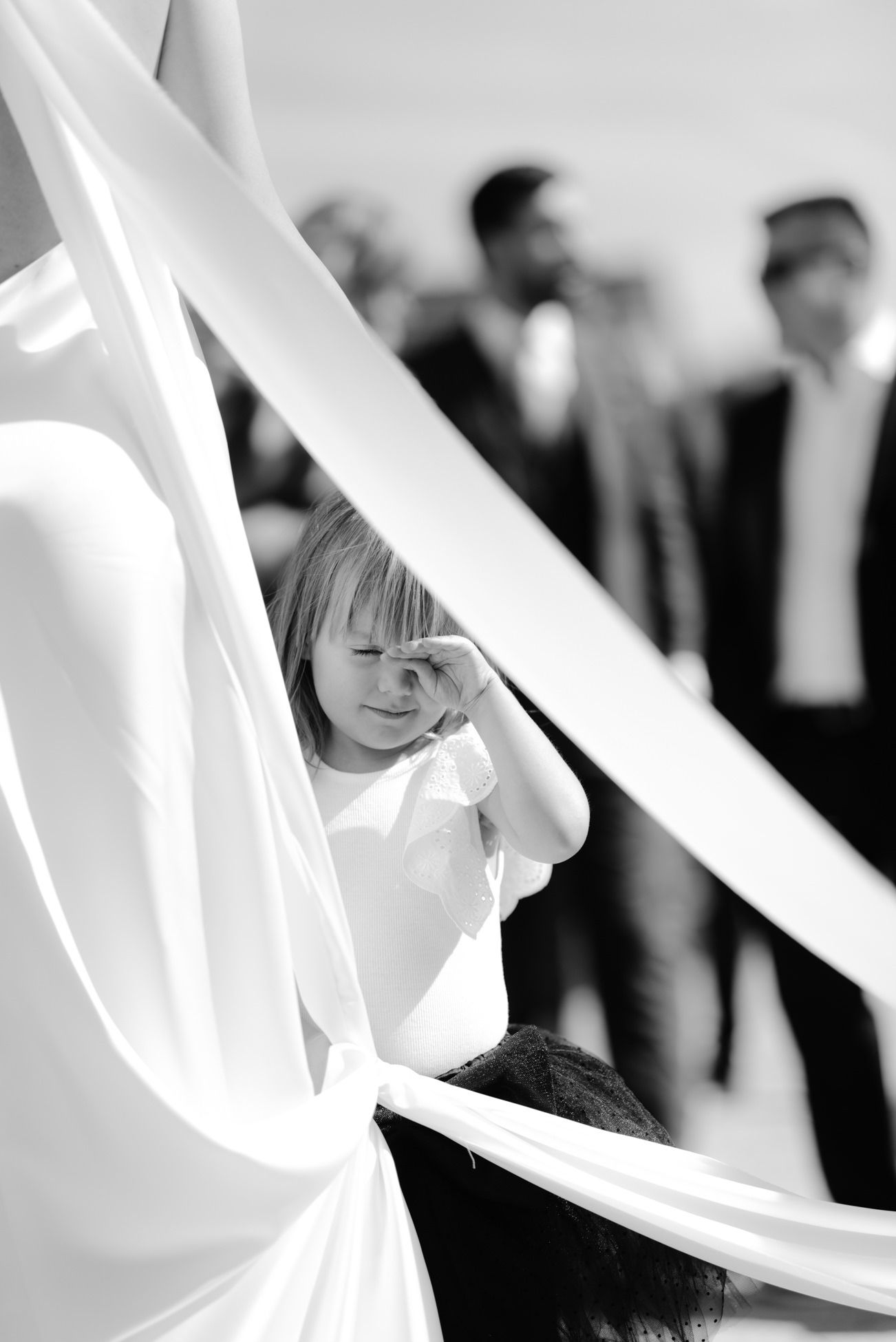 A child peeks playfully through flowing white fabric at a formal event while people stand in the background.
