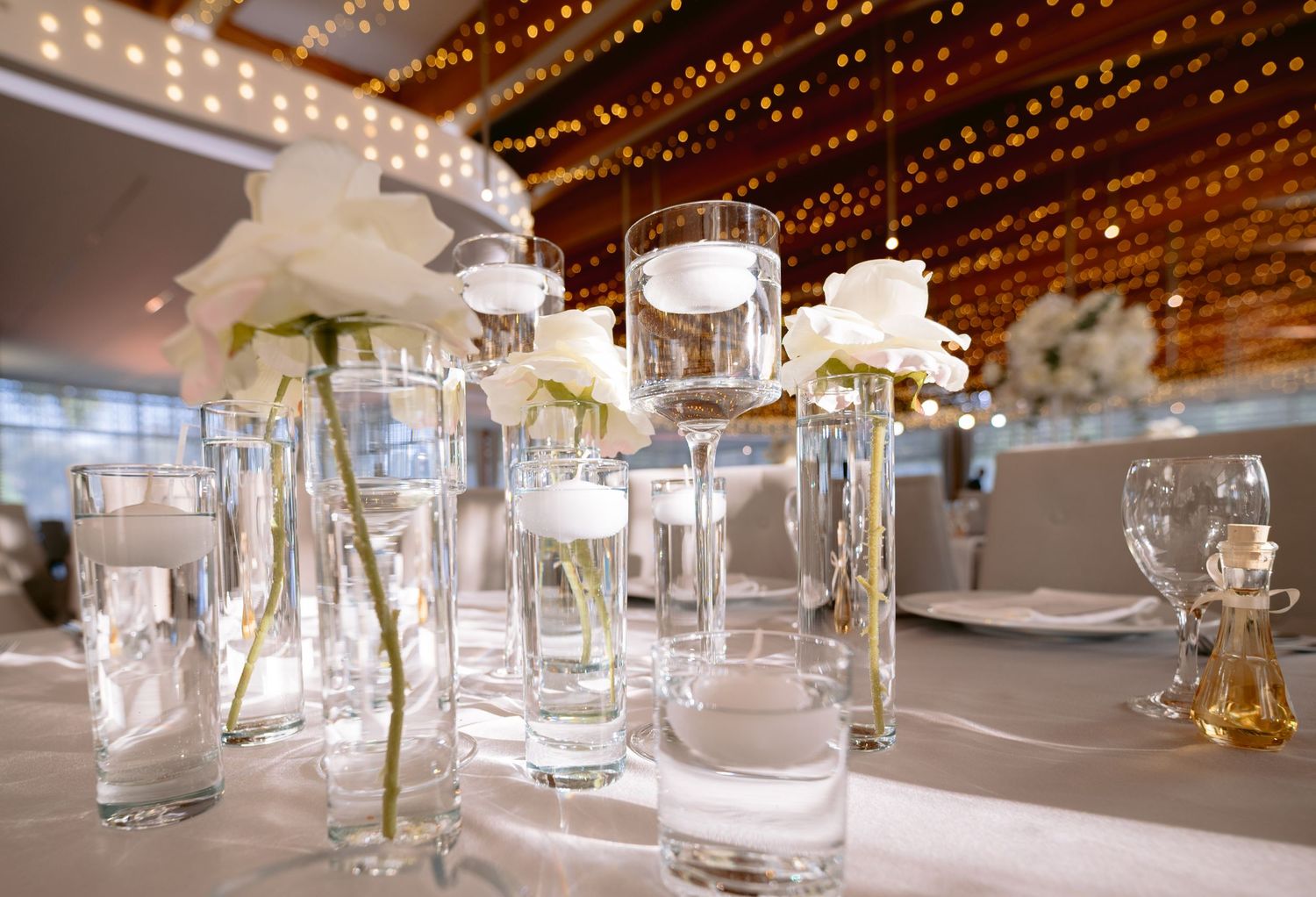 Elegant wedding centerpiece with white roses in tall glass vases illuminated by warm string lights overhead.