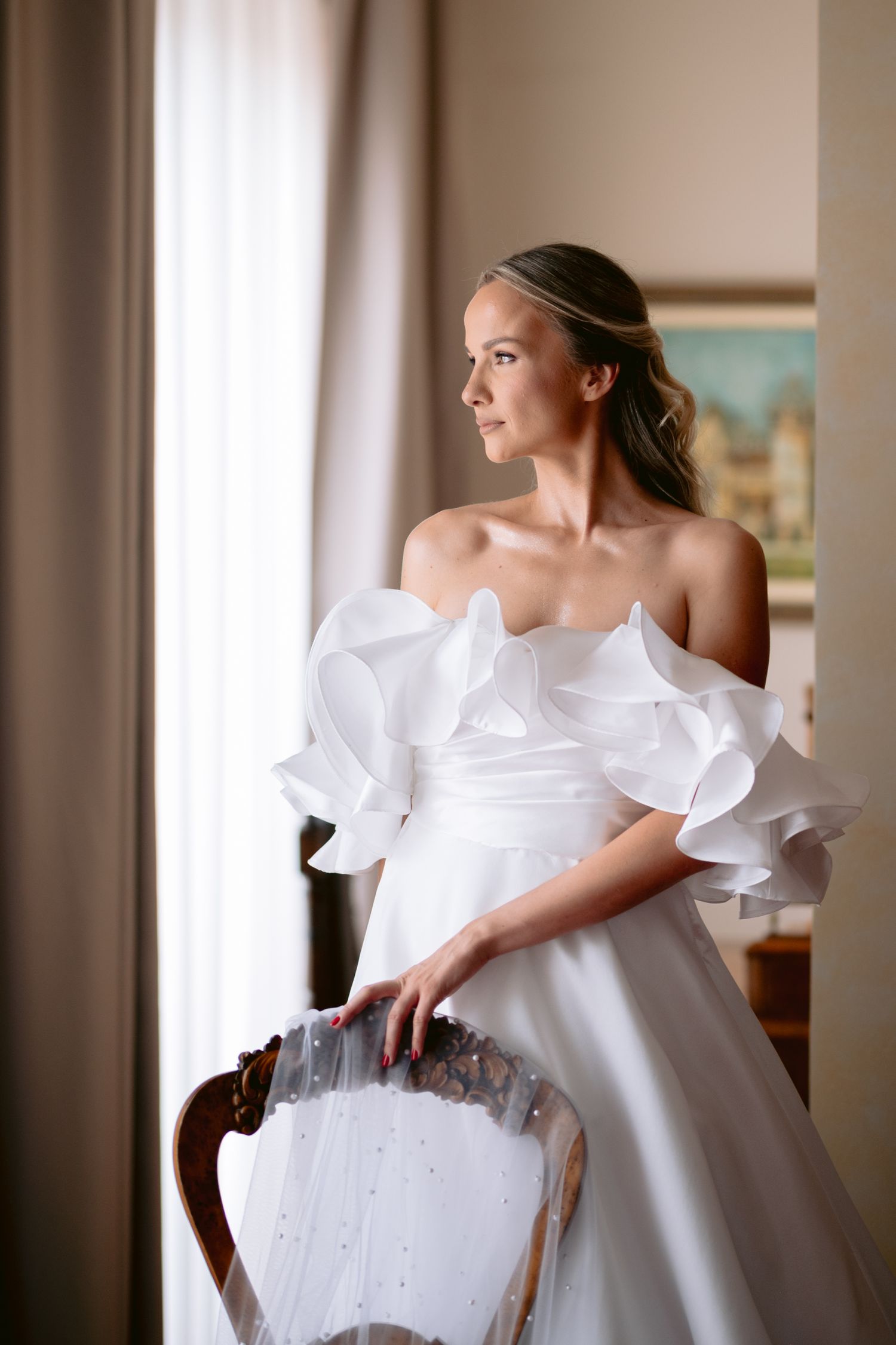 Bride in an elegant white wedding gown with ruffled details sits gracefully in a vintage chair.