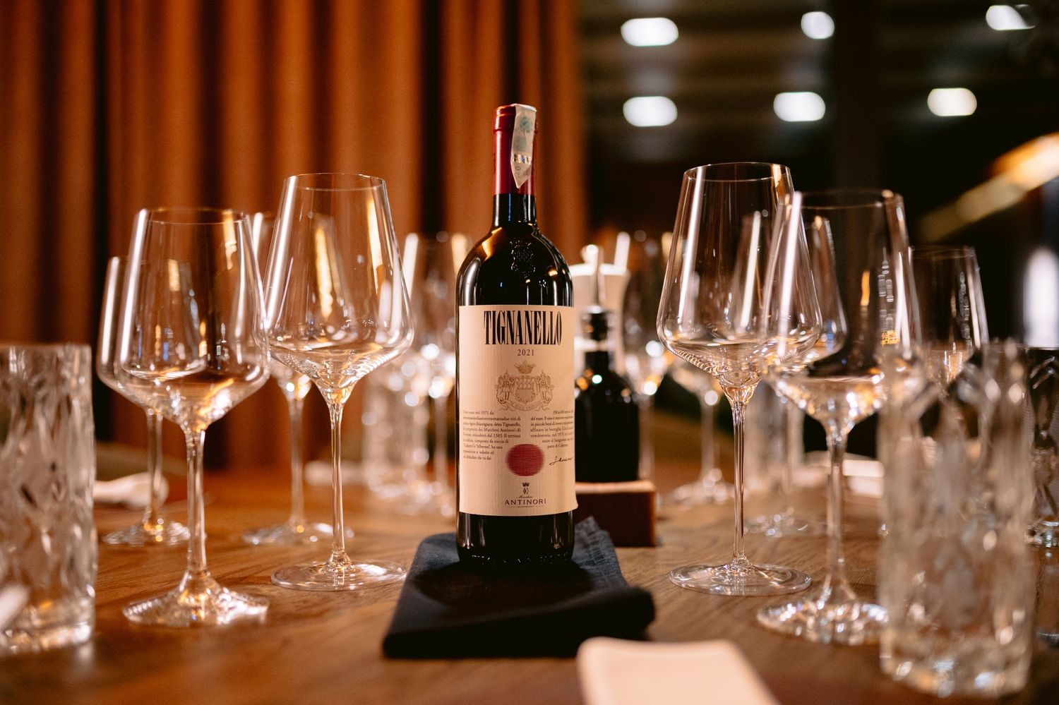 Wine bottles and empty glasses line a wooden bar counter under warm lighting in an upscale restaurant setting.