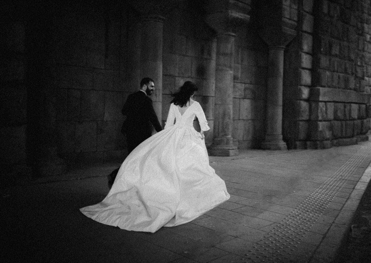 A bride in a flowing white wedding dress runs through a dark stone archway in black and white photography.
