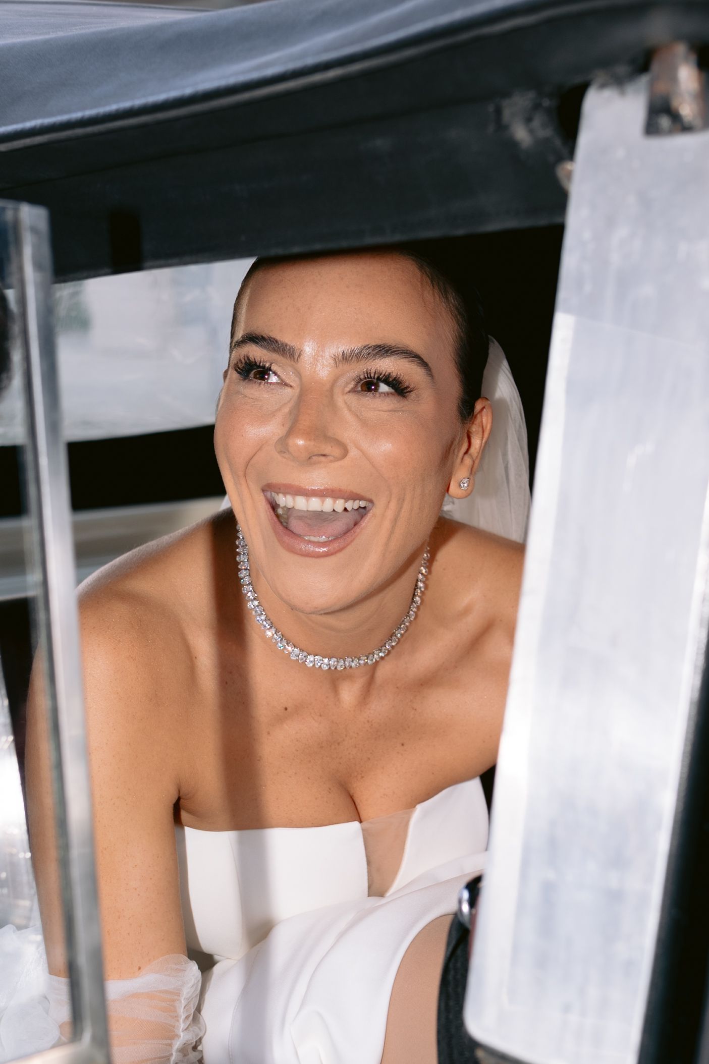 A bride wearing a white strapless dress and diamond necklace smiles joyfully while sitting in a vehicle.