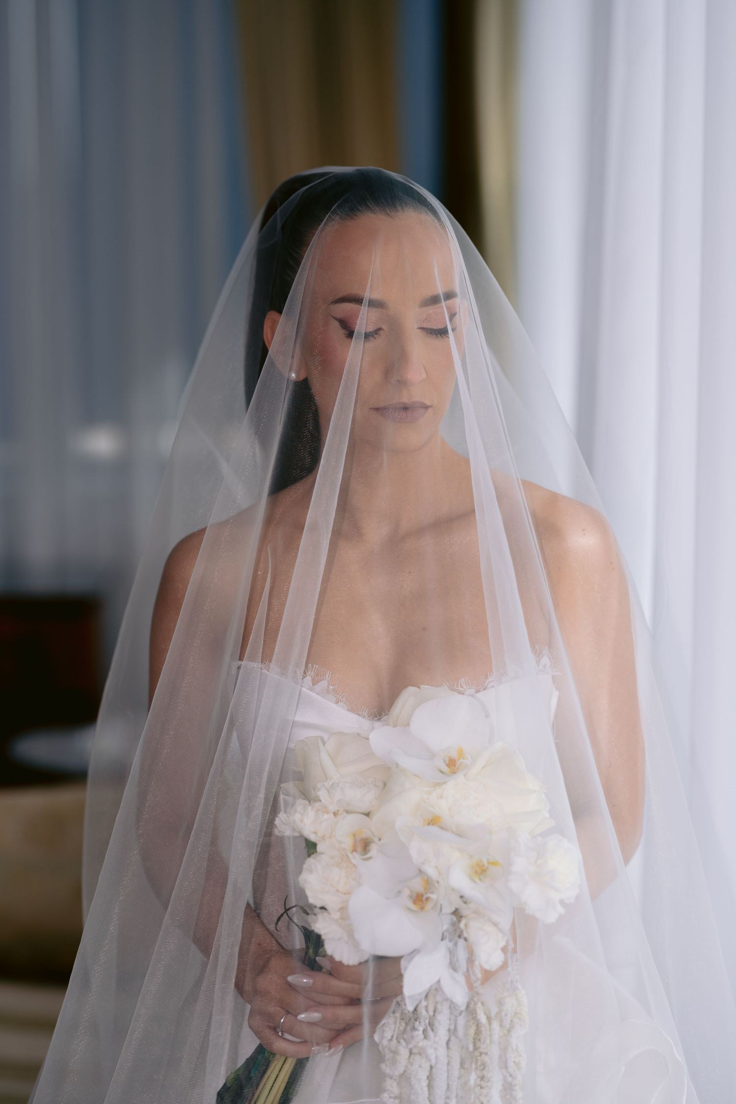 A bride in a white wedding dress and veil stands elegantly in a softly lit indoor setting.