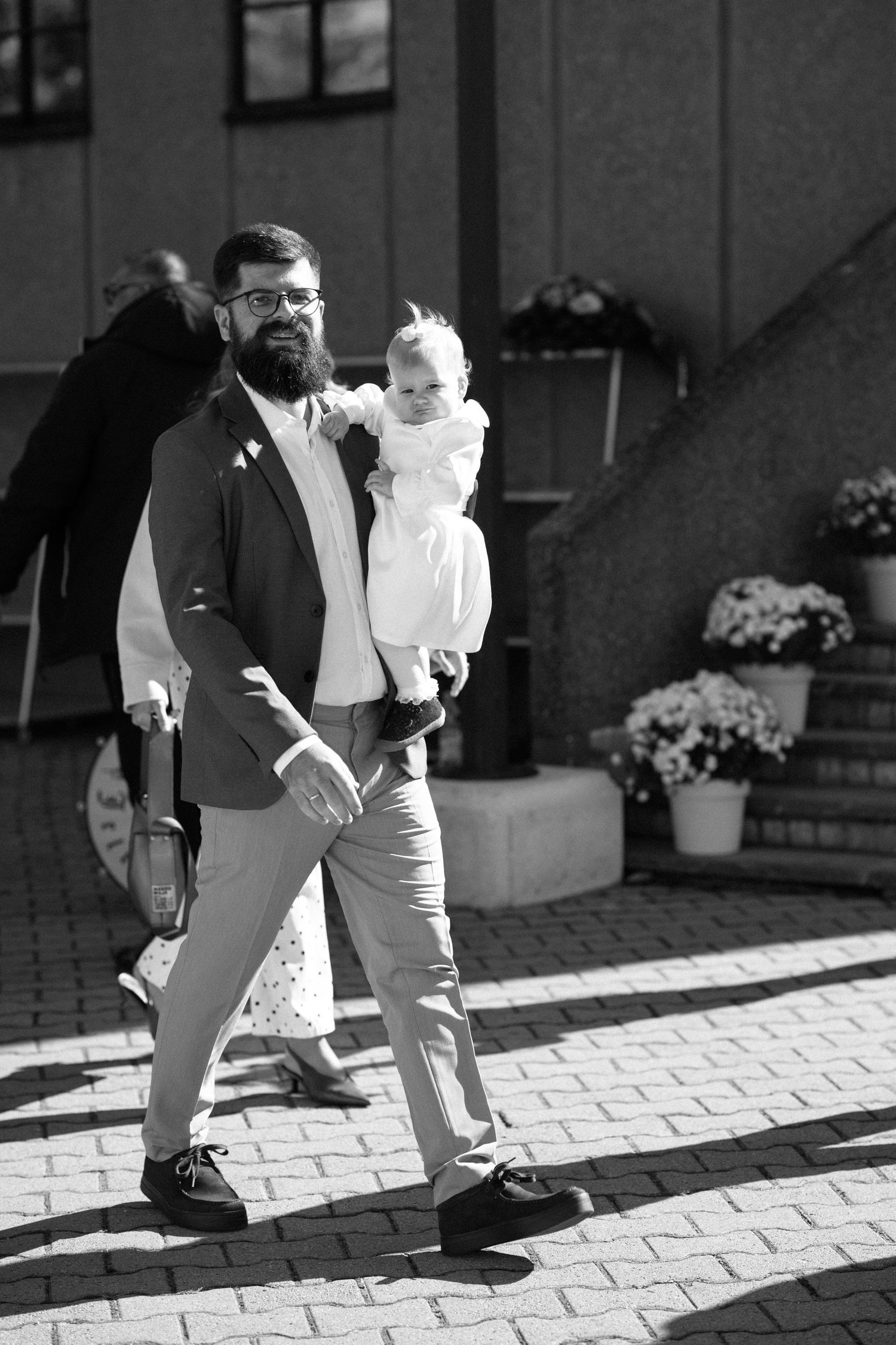 A bearded man in glasses and a blazer walks down a sunlit brick path carrying a smiling baby in a white dress, with flower pots nearby.